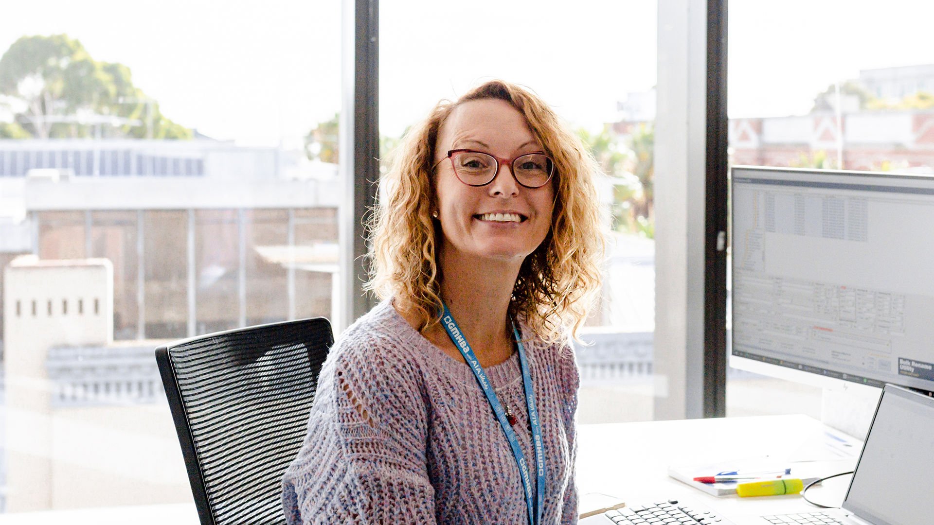 Female with glasses and curly hair sitting at office desk with large windows behind